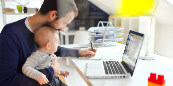 Dad holding baby while working at a desk in front of a laptop